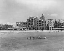 Load image into Gallery viewer, Black and white photograph of the Royal Hawaiian Hotel on Waikiki Beach taken from the water. Beachgoers in the sand and water. An outrigger canoe with passengers in the foreground.