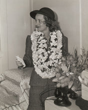 Load image into Gallery viewer, Black and white photograph of actress Bette Davis sitting on a bed of the Matson Lines cruise ship, Lurline, wearing many flower leis.
