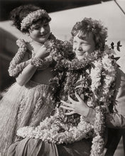 Load image into Gallery viewer, Black and white photograph of a young girl standing next to a sitting Amelia Earhart wearing multiple flower leis.