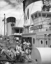 Load image into Gallery viewer, Black and white photograph of the ship deck of the Matsonia featuring 4 hula dancers, 3 musicians, and 2 crew members, all wearing flower leis.