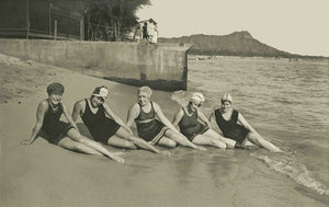 Grey sepia toned photograph of five women in early 1900's swimsuits posed in a half-reclining position on Waikiki Beach shoreline. Diamond Head is seen in the background.