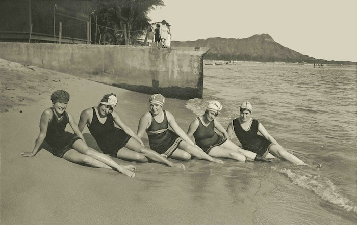 Grey sepia toned photograph of five women in early 1900's swimsuits posed in a half-reclining position on Waikiki Beach shoreline. Diamond Head is seen in the background.