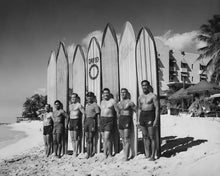 Load image into Gallery viewer, Group of surfers with their surfboards on a beach, with a building and palm trees in the background.