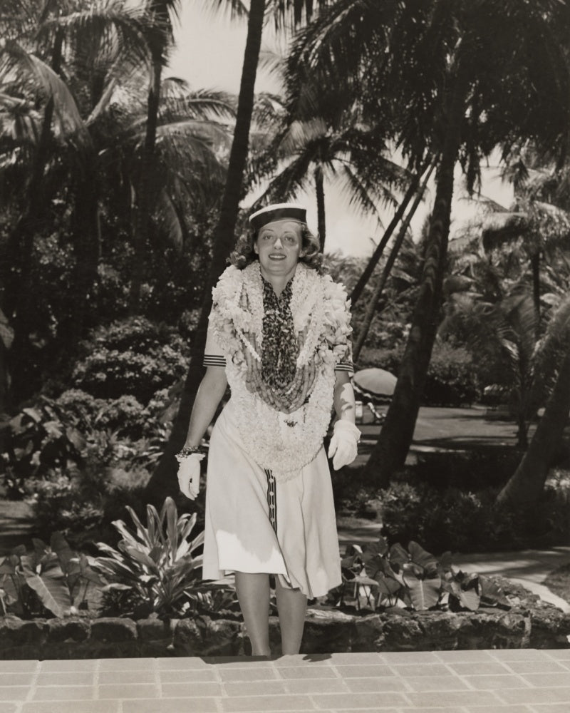 Black and white vintage photo of young Bette Davis wearing multiple lei with a tropical setting behind her. 