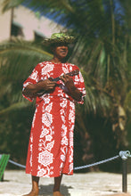 Load image into Gallery viewer, A man wearing a red and white floral print dress and a green straw hat, holding a ukulele outdoors with palm trees in the background.
