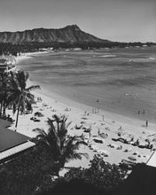 Load image into Gallery viewer, Black and white photograph of a mountain peak overlooking a beach and and bay. There are palm trees, umbrellas, and beach bathers on the sand.