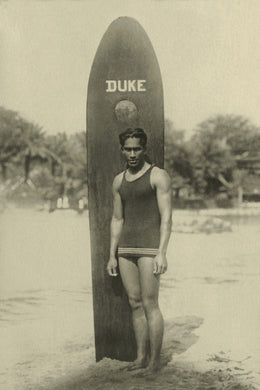 Photograph in light brown sepia tones of famous swimmer and surfer from Hawaii, Duke Kahanamoku standing in front of a sufrboard with 