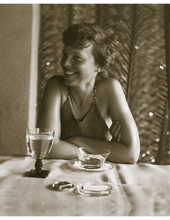 Load image into Gallery viewer, Brown sepia toned photograph of Amelia Earhart in a swimsuit smiling while sitting at a table. There is a glass of water, cup of tea, matches and an ashtray with cigarettes on the table.