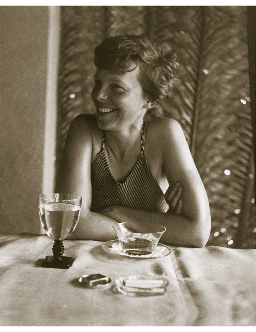 Brown sepia toned photograph of Amelia Earhart in a swimsuit smiling while sitting at a table. There is a glass of water, cup of tea, matches and an ashtray with cigarettes on the table.