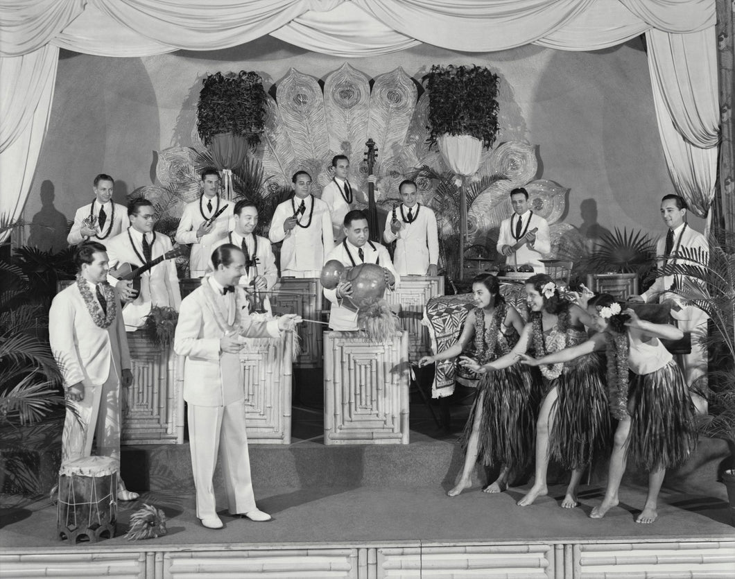 Black and white photo of bandleader Harry Owens leading the Royal Hawaiian Orchestra and accompanied by female hula dancers.