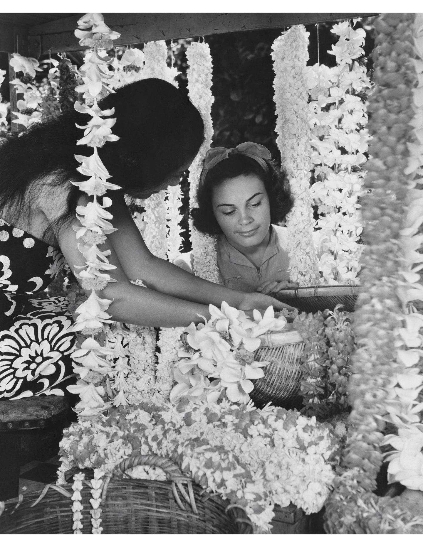 Black and white photograph of a woman working at a lei stand assisting a female customer.