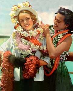 Color photograph of a woman adorned with many flower lei and a woman on the right also wearing multiple lei fixing flowers on the other woman.