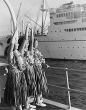 Load image into Gallery viewer, Black and white photograph of three women in hula-style grass skirts and flower leis standing on a ship deck, waving toward a large ocean liner in the harbor.