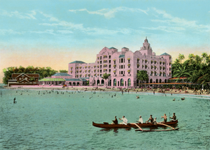Colorful vintage photograph of the pink Royal Hawaiian Hotel as viewed from the ocean with water and outigger canoe with paddlers in foreground and the beach and hotel in the back.