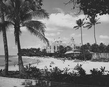 Load image into Gallery viewer, Black and white photo of a beach view from between palm trees facing a hotel. 