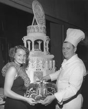 Load image into Gallery viewer, Black-and-white photo of two people holding a tray of food, standing in front of an intricately decorated cake.