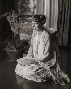 Black and white photograph of Amelia Earhart wearing a silk kimono robe while sitting with a book in her lap and giving the camera her profile as she looks in the distance.