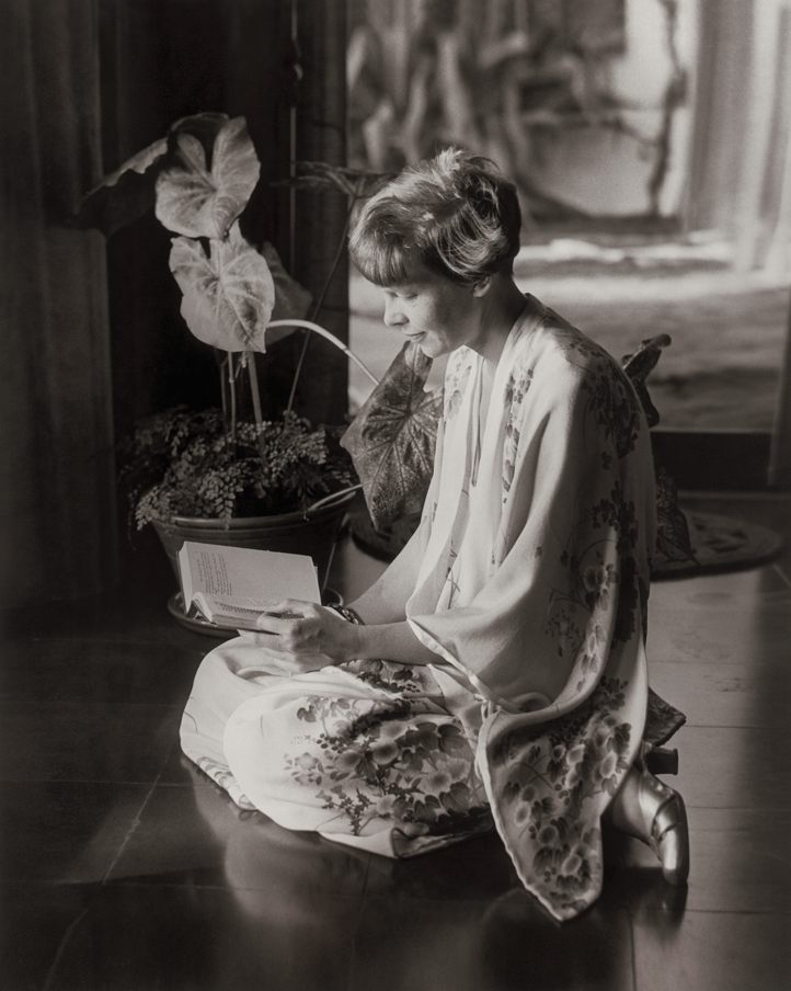 Black and white photograph of Amelia Earhart wearing a kimono robe, sitting on the floor, reading a book.