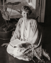 Load image into Gallery viewer, Black and white photograph of Amelia Earhart wearing a silk kimono robe while sitting on the floor with a book in her lap and smiling at the camera.