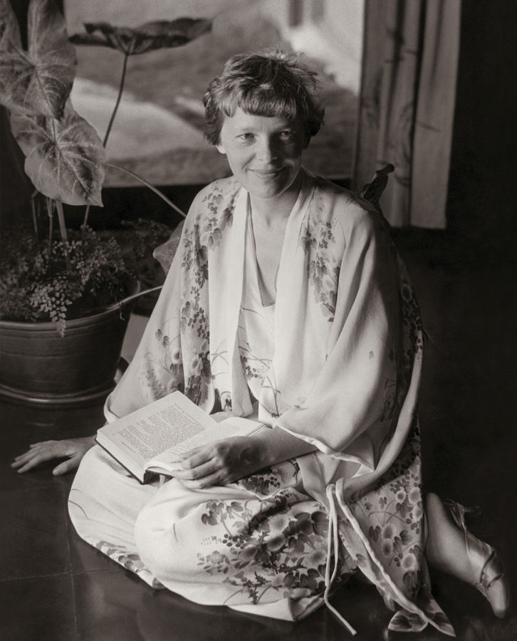 Black and white photograph of Amelia Earhart wearing a silk kimono robe while sitting on the floor with a book in her lap and smiling at the camera.