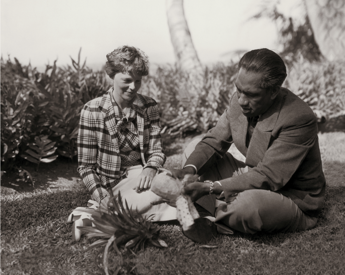 Black and white photograph of Amelia Earhart shares a freshly peeled pineapple while sitting on the grass with Duke Kahanamoku. 