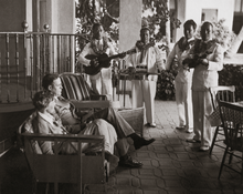 Load image into Gallery viewer, Black and white photograph of Amelia Earhart sits with her husband on a patio while four musicians in white suits and flower leis play various instruments.