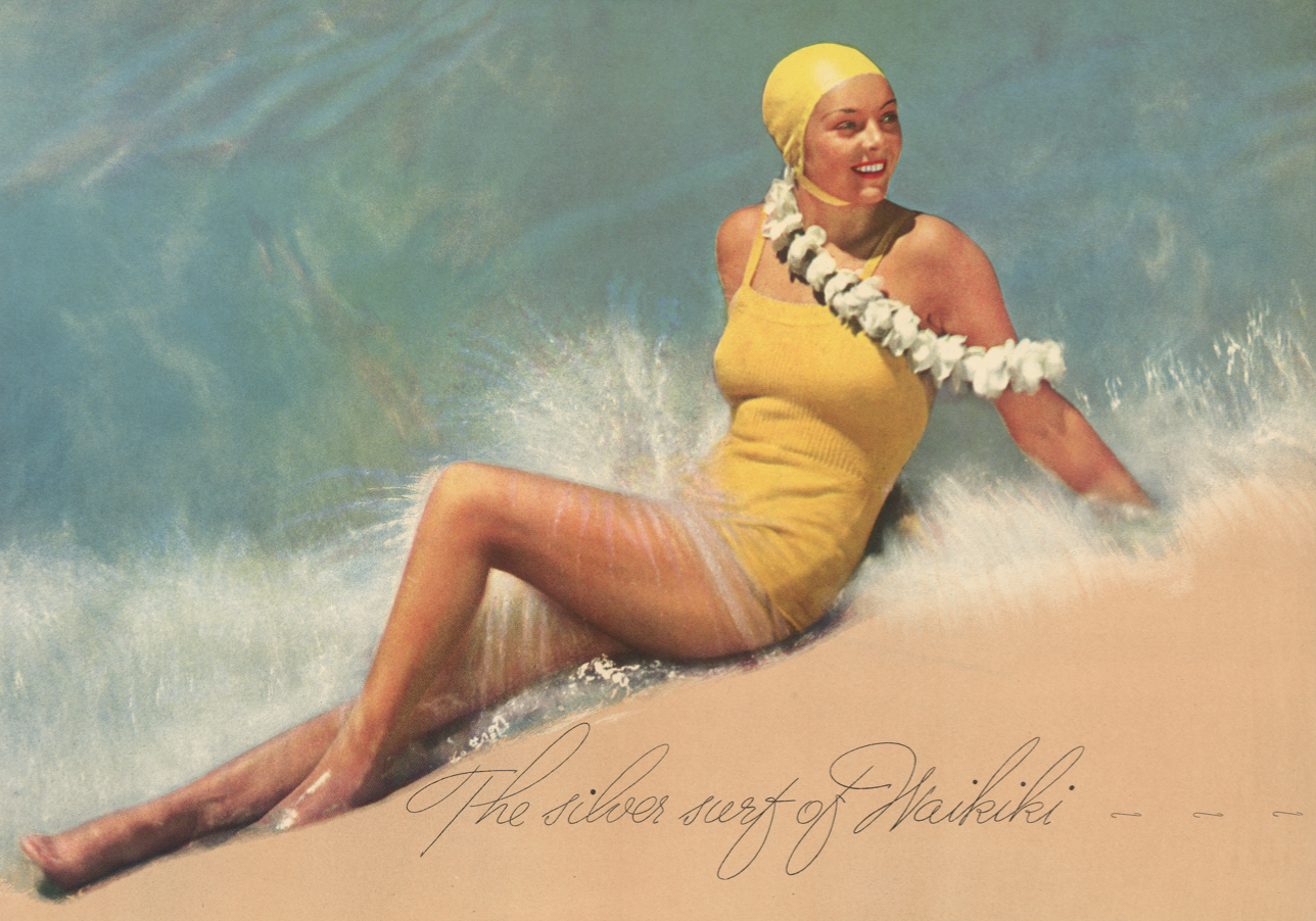 Color photograph of a woman in yellow swimsuit and yellow bathing cap reclining on a beach with the water behind her and the sand in front. 
