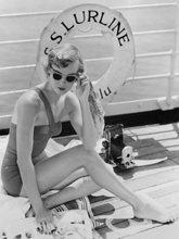 Load image into Gallery viewer, Black and white vintage photograph of a woman in a swimsuit wearing sunglasses and sitting on a ship deck. There is a camera behind her and a lifesaver with S.S. Lurline written on it.