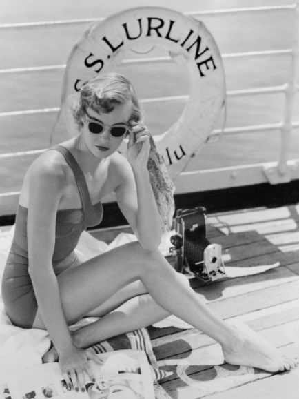 Black and white vintage photograph of a woman in a swimsuit wearing sunglasses and sitting on a ship deck. There is a camera behind her and a lifesaver with S.S. Lurline written on it.
