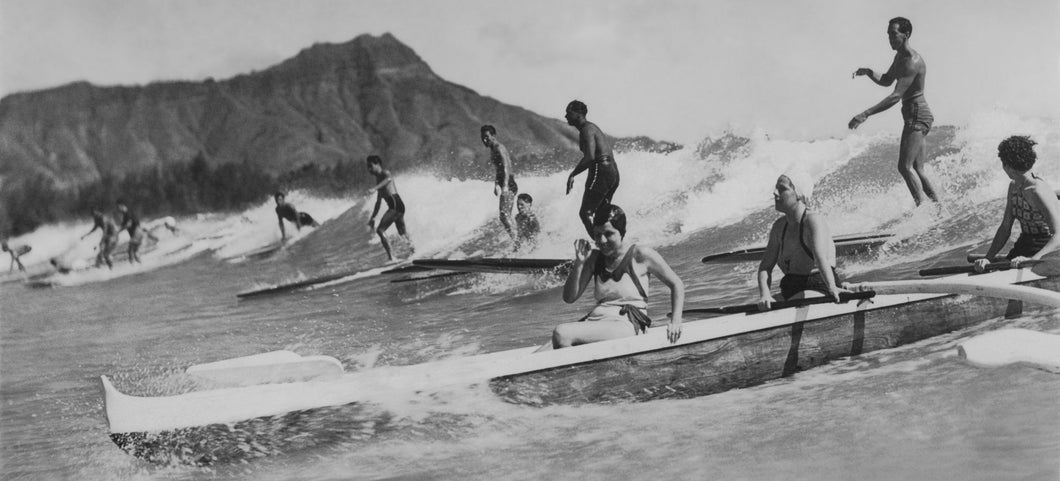 Black and white vintage photo of surfers riding a wave alongside an outrigger canoe in Waikiki, Hawaii. Diamond Head crater in the background 