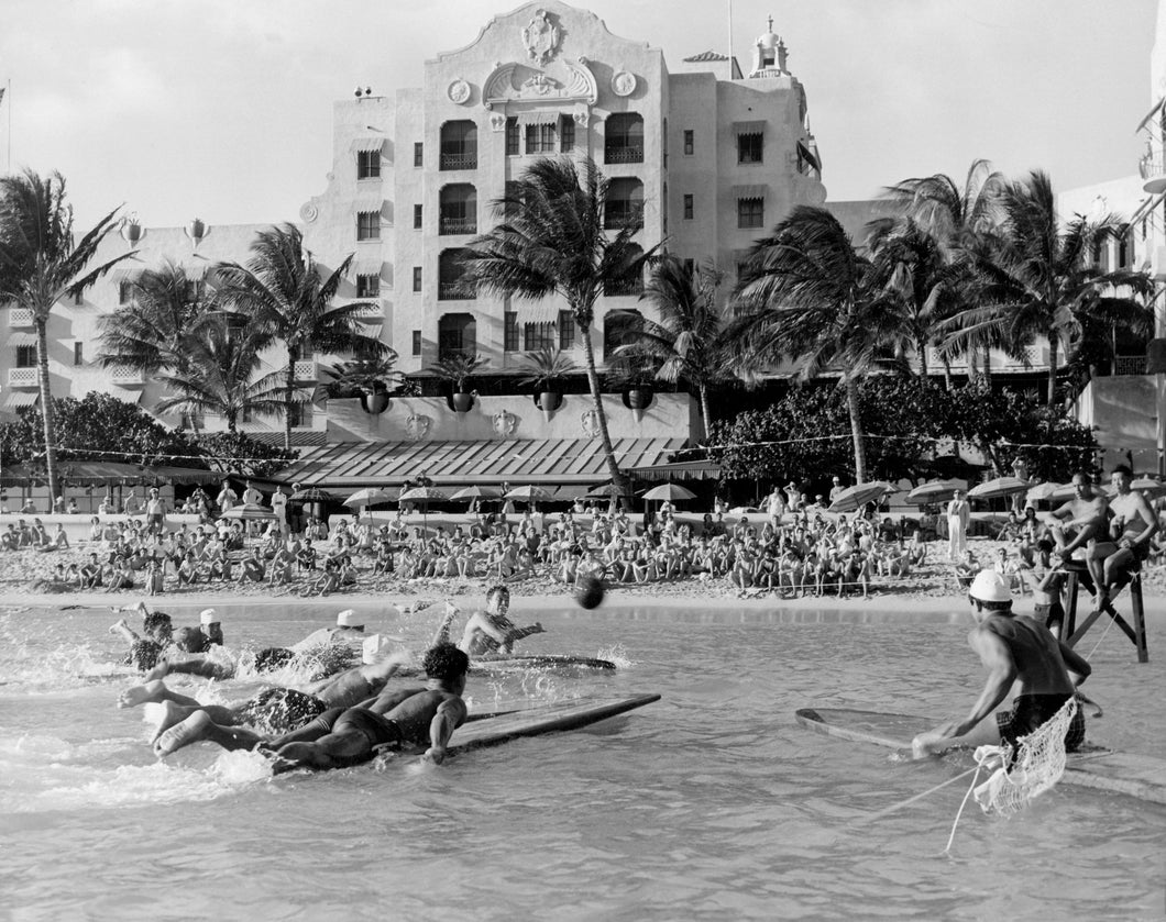 Black and white photograph of people lying on surfboards and playing water polo in front of the Royal Hawaiian Hotel while onlookers sit on the sand.