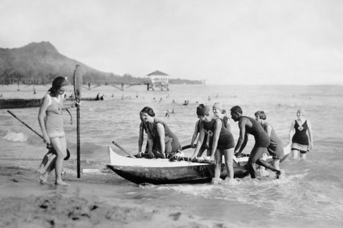 Black and white photo of women pushing a canoe onto Waikiki Beach with the help of David Kahanamoku. 
