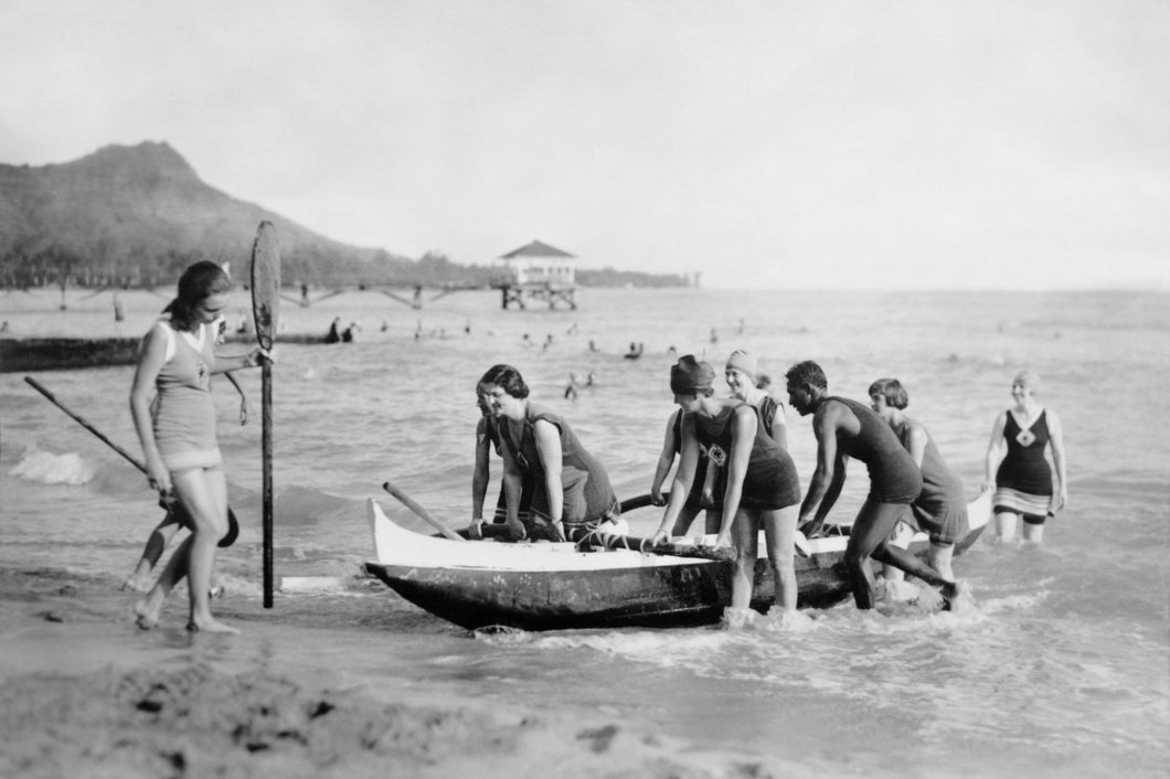 Black and white photo of women pushing a canoe onto Waikiki Beach with the help of David Kahanamoku. 