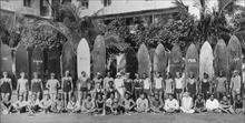 Load image into Gallery viewer, Black and white panoramic photo of tall surfboards lined up in the back, a row of men standing in front of them, and a row of men sitting on the ground in front of the standing men. 