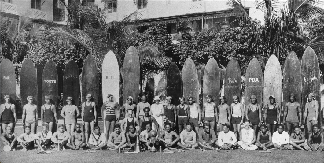 Black and white panoramic photo of tall surfboards lined up in the back, a row of men standing in front of them, and a row of men sitting on the ground in front of the standing men. 