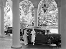 Load image into Gallery viewer, Historical black and white 1930s era photograph of a woman stepping out of a car parked beneath tall columned arches, while the door is being held open by a hotel bellman.