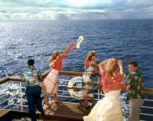 Load image into Gallery viewer, Color photograph of a 3 women and 2 men standing on the deck of a cruise ship facing the open ocean. One woman wearing a red top and red and white skirt is throwing a white lei into the sea. 