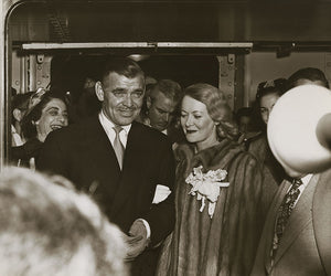 Black and white photograph of actor Clark Gable in a suit accompanying Lady Sylvia Ashley who is wearing a fur coat among a crowd of people on the Matson Lines cruise ship, Lurline.