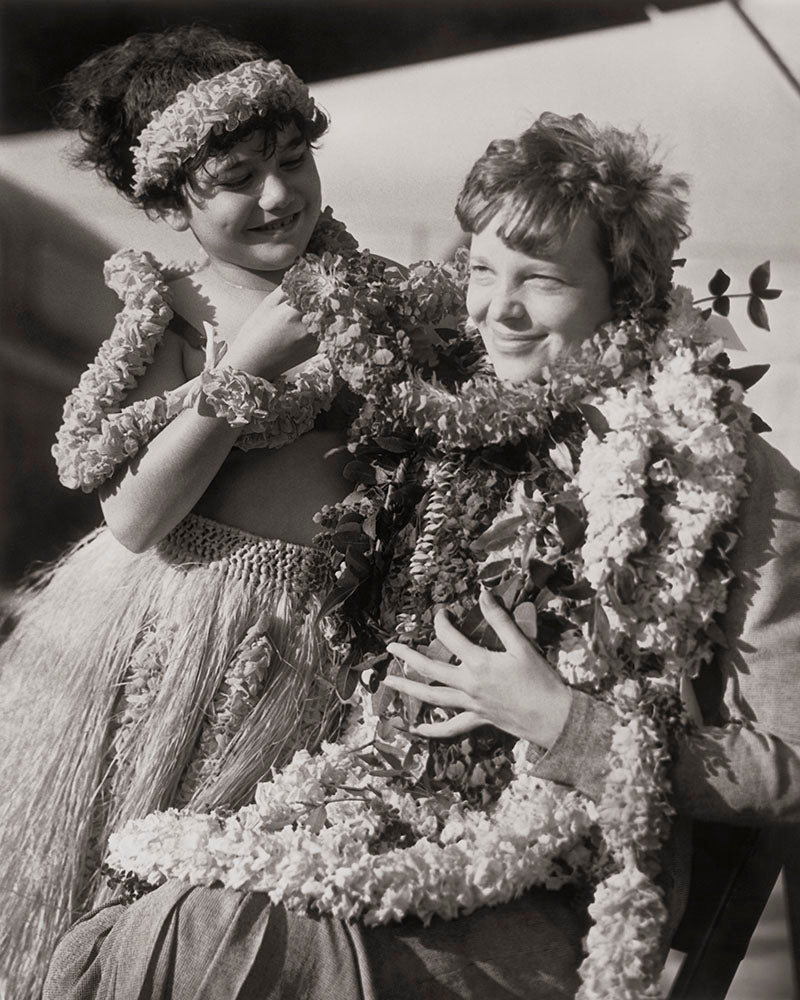Black and white photograph of a young girl standing next to a sitting Amelia Earhart wearing multiple flower leis.