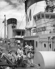 Load image into Gallery viewer, Black and white photograph of the ship deck of the Matsonia featuring 4 hula dancers, 3 musicians, and 2 crew members, all wearing flower leis.  