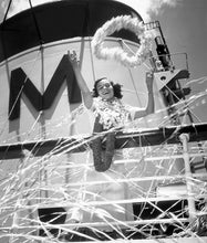 Load image into Gallery viewer, Black and white photograph of a woman wearing multiple flower leis and standing on the top deck of a Matson Line cruise ship tossing a flower lei in the air. The Matson &quot;M&quot; is on the smokestack behind her.