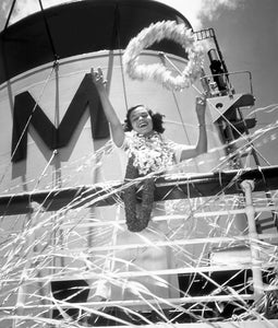 Black and white photograph of a woman wearing multiple flower leis and standing on the top deck of a Matson Line cruise ship tossing a flower lei in the air. The Matson "M" is on the smokestack behind her.