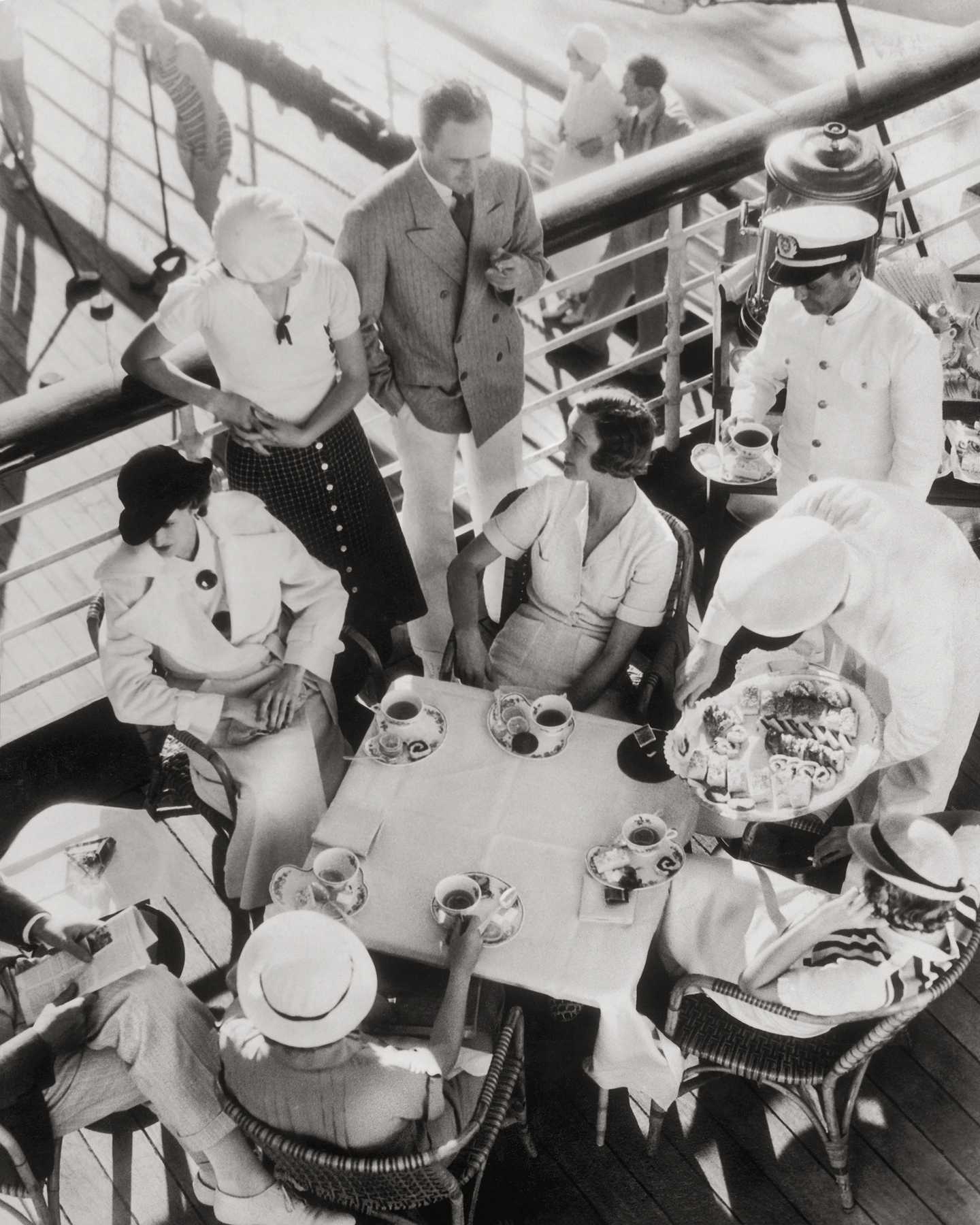 Black and white photograph of a group of people in the 1930s on the deck of a ship sitting at a table having tea. By photographer Edward Steichen.