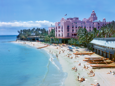 Full color photograph of the pink Royal Hawaiian Hotel and Outrigger Canoe Club on the right and the ocean on the left with Waikiki Beach in the center. Many bathers and outrigger canoes are lined up on the sand..