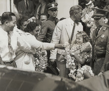 Load image into Gallery viewer, Black and white photograph of a young Shirley Temple wearing multiple flower leis being escorted by police among a crowd while a woman hands a lei to her.