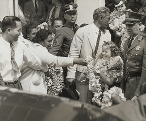Black and white photograph of a young Shirley Temple wearing multiple flower leis being escorted by police among a crowd while a woman hands a lei to her.