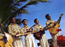 Load image into Gallery viewer, Color photograph of musicians and playing their instruments outdoors. 5 instruments and 4 musicians are pictured. 4 men wearing white pants and matching patterned shirts and leis under a blue sky with a palm tree in background to the left.